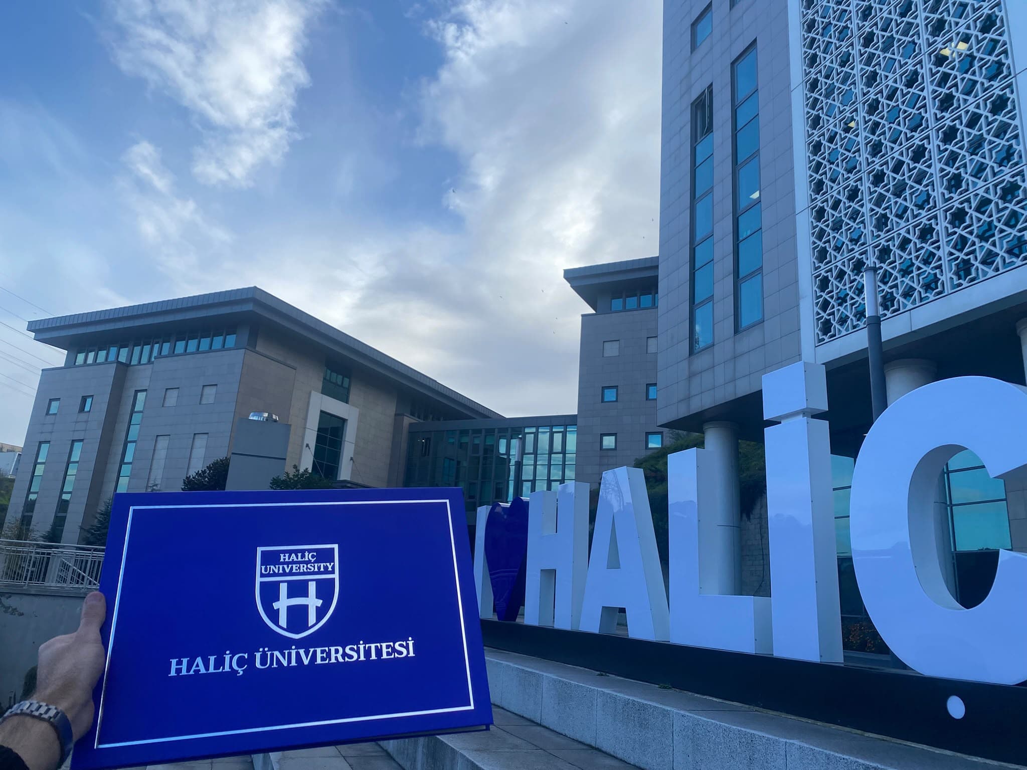 A closed Haliç University diploma folder in front of the university campus